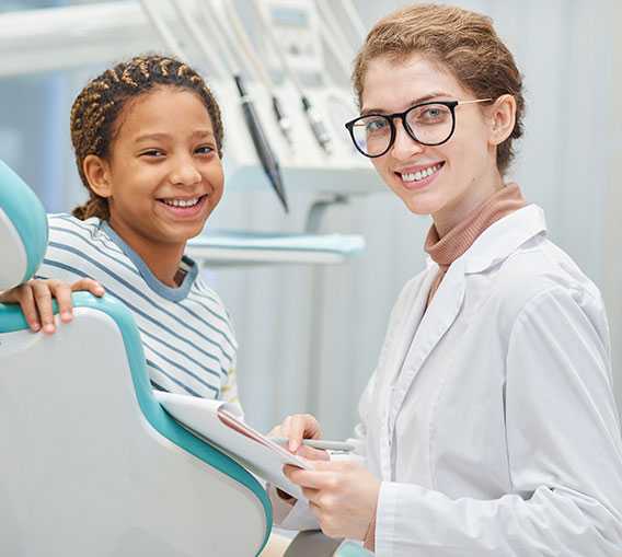 A smiling young patient and a dental professional with a clipboard are posing in a dental clinic.